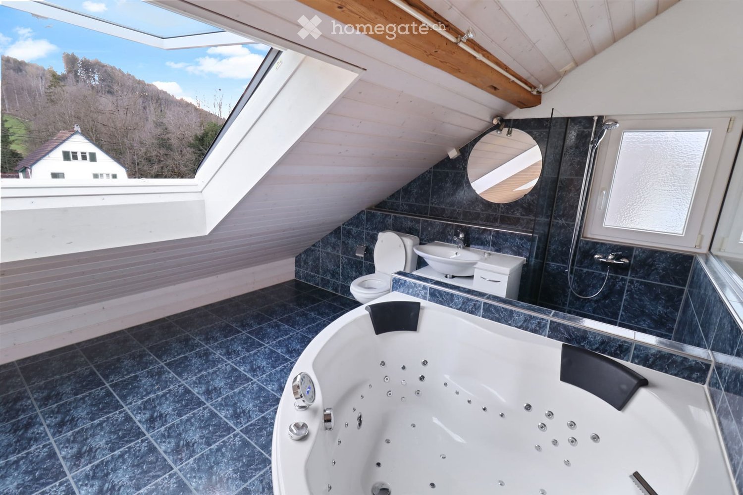 Modern bathroom with a large bathtub, toilet, sink, and shower, featuring dark tiles and a skylight window offering a scenic view.