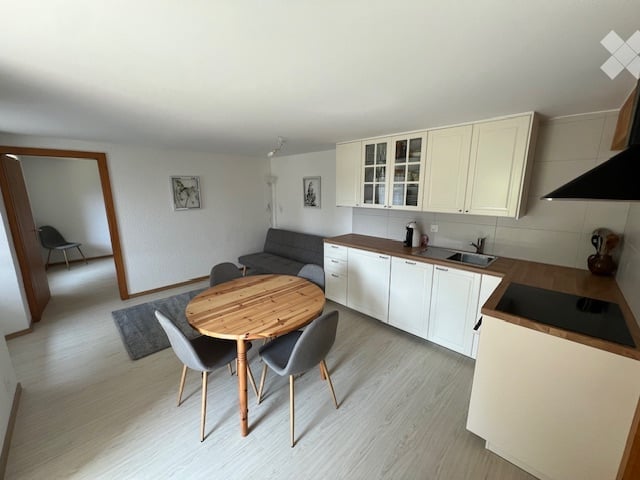 Open-plan living and dining area with a grey sofa, wooden dining table with four chairs, white kitchen cabinets, sink, and stove. The space features light wood flooring and white walls.