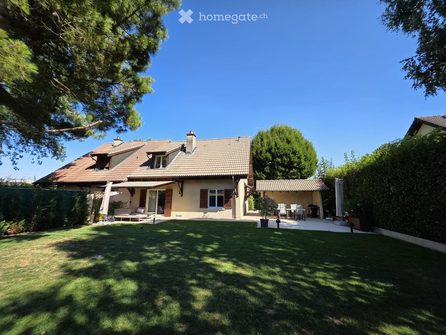 Two-story house with a tiled roof, surrounded by a well-maintained lawn and trees. Features include a covered patio area with outdoor furniture and a barbecue.
