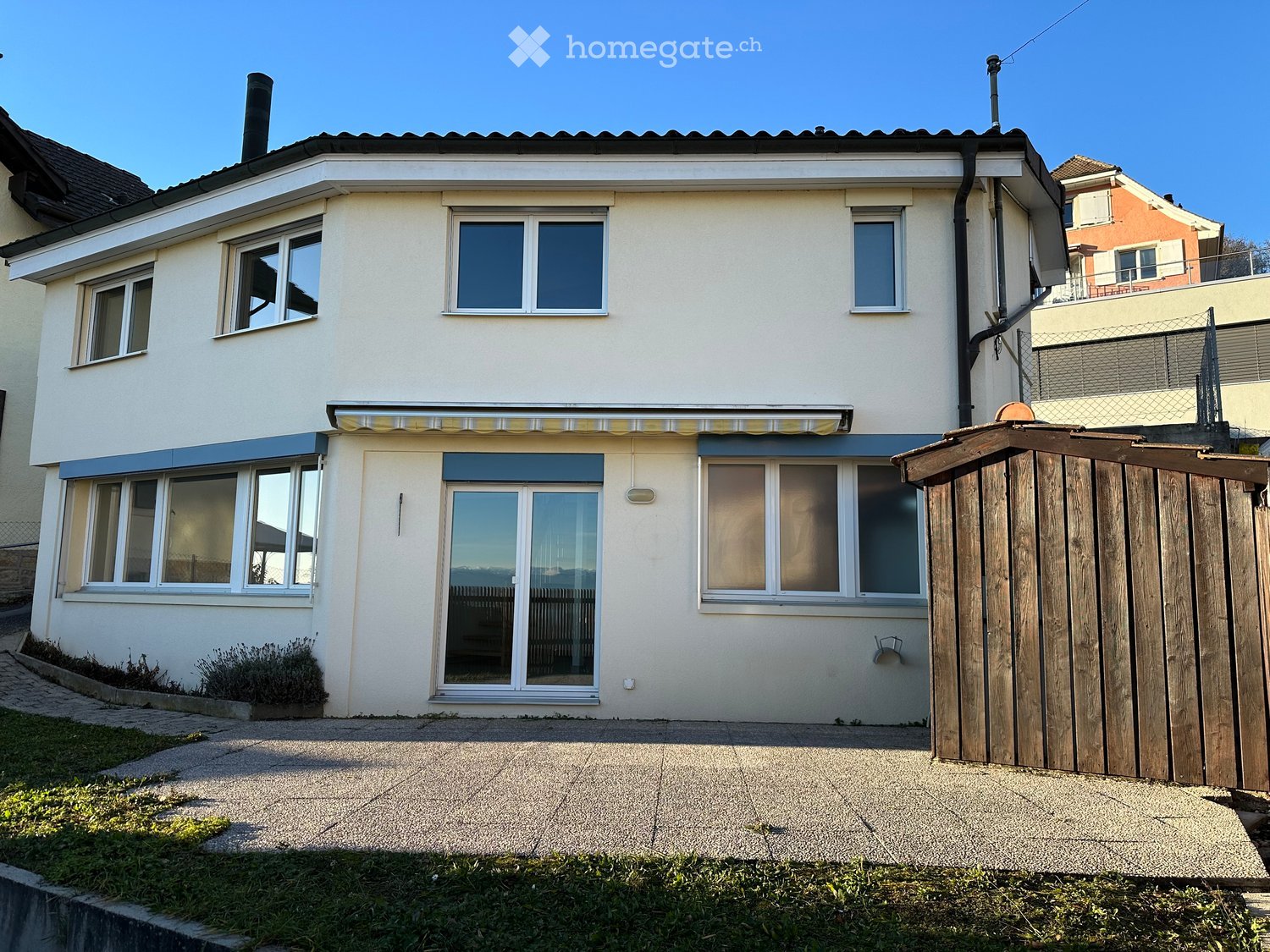 Two-story house with light-colored exterior, multiple windows, and a covered entrance area. Features a wooden fence and small garden area.