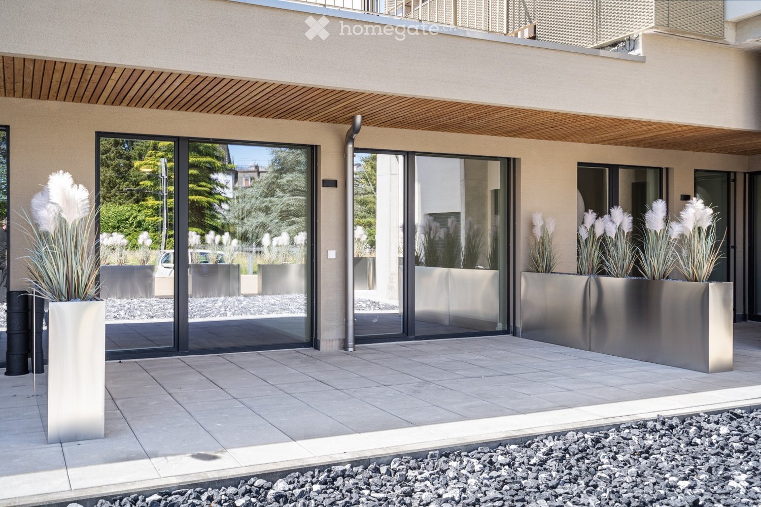 Modern outdoor area with large glass sliding doors, tiled floor, and tall metallic planters with white decorative plants.