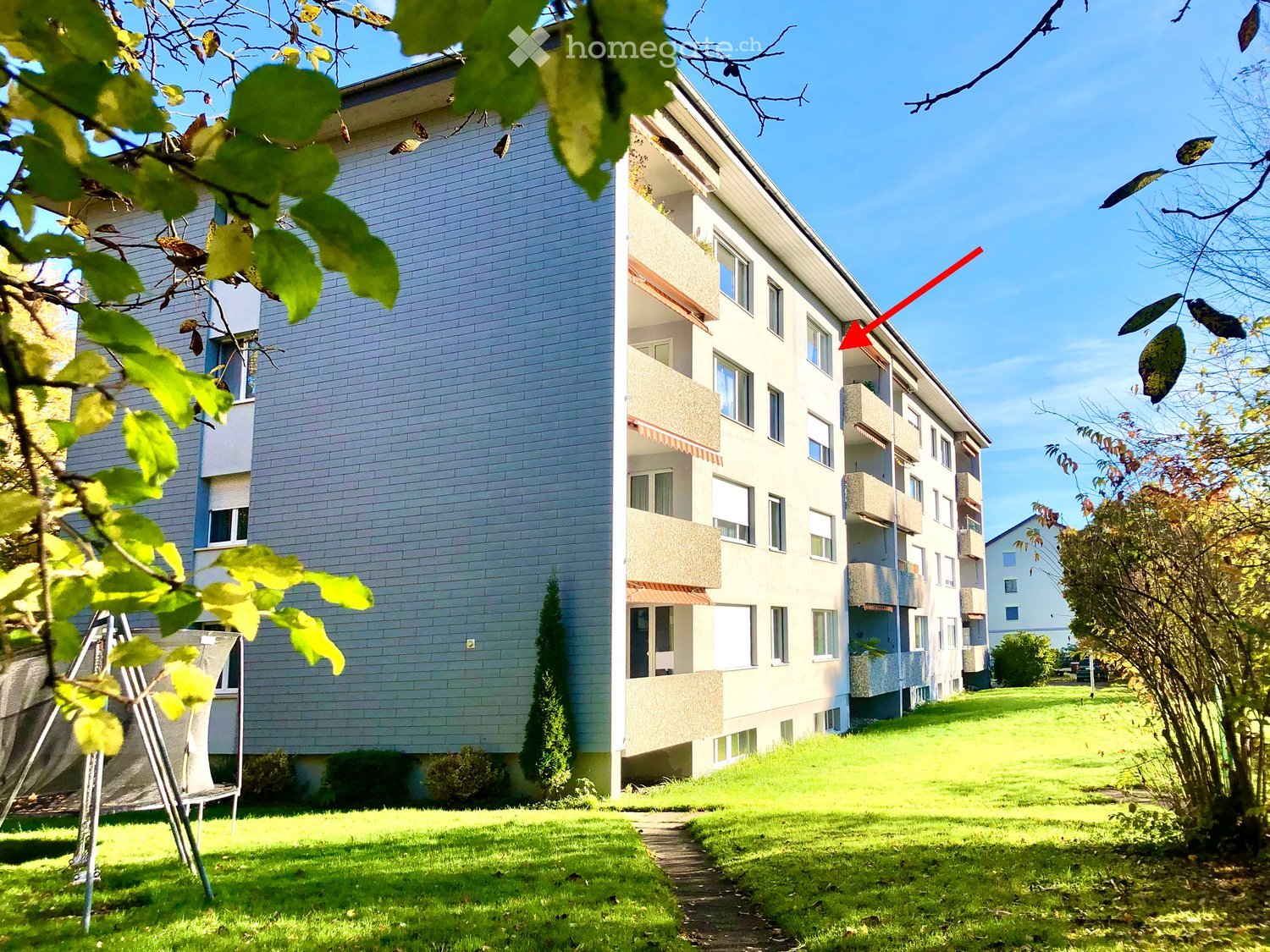 Multi-story residential building with balconies, surrounded by green lawn and trees.