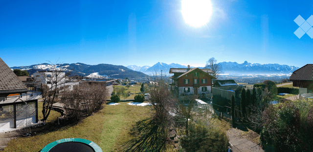 A panoramic view of a residential area with houses, green lawns, trees, and distant snow-capped mountains under a bright blue sky.