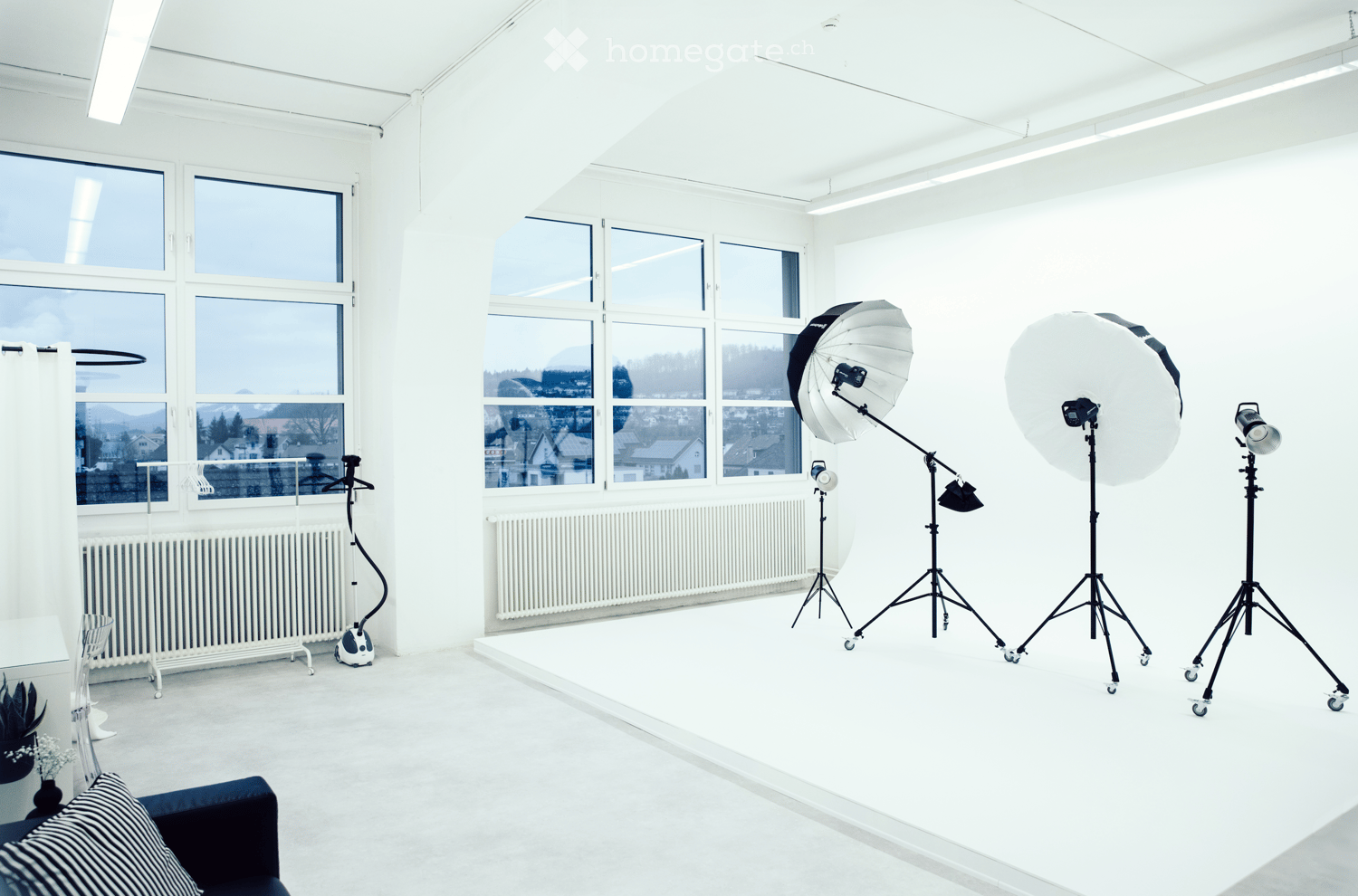 Bright, minimalist room with large windows, white walls, and a sofa. Includes a small table, chair, radiator, curtain, plant, and photography equipment such as tripods, umbrellas, and cameras.