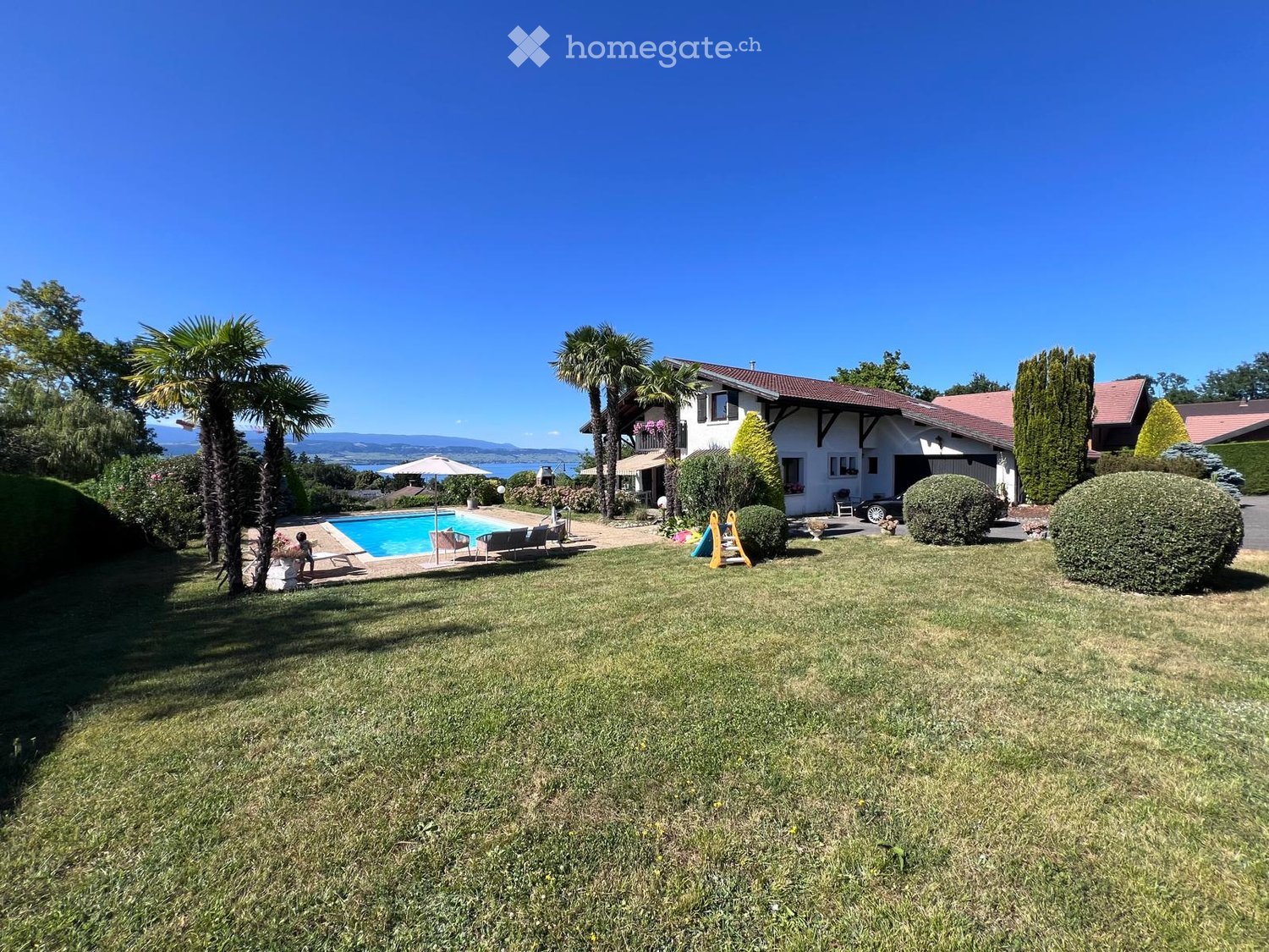 A well-maintained garden with a swimming pool, lounge chairs, and a large house with a garage and balcony.