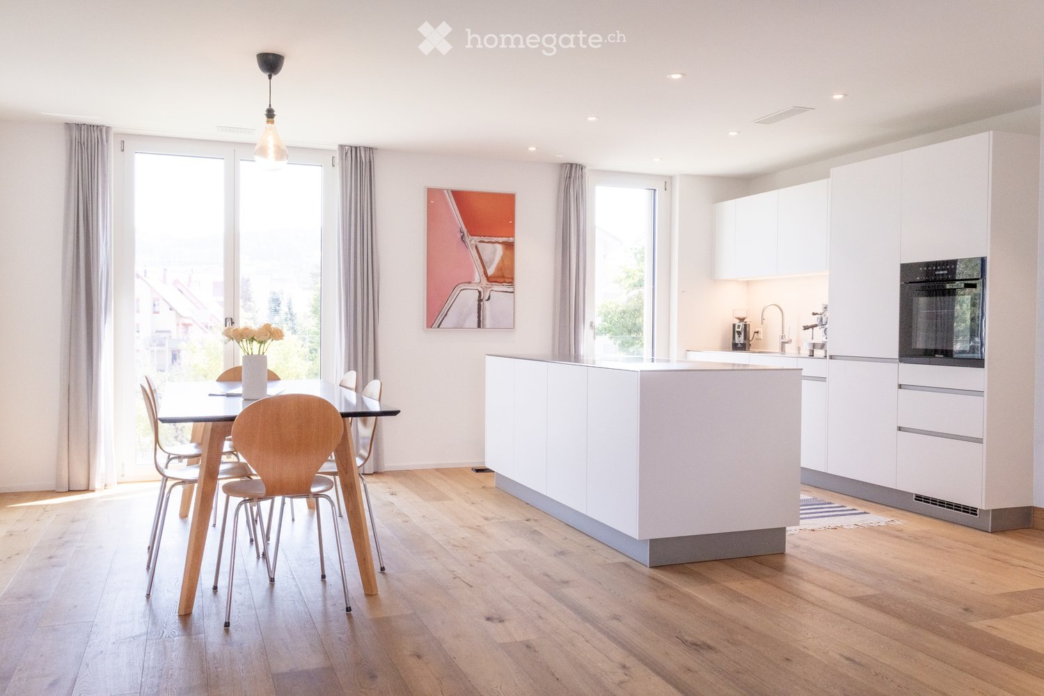 Modern kitchen and dining area with white cabinets, a central island, a dining table with chairs, and a vase of flowers on the table. The space features wooden flooring and large windows with curtains.