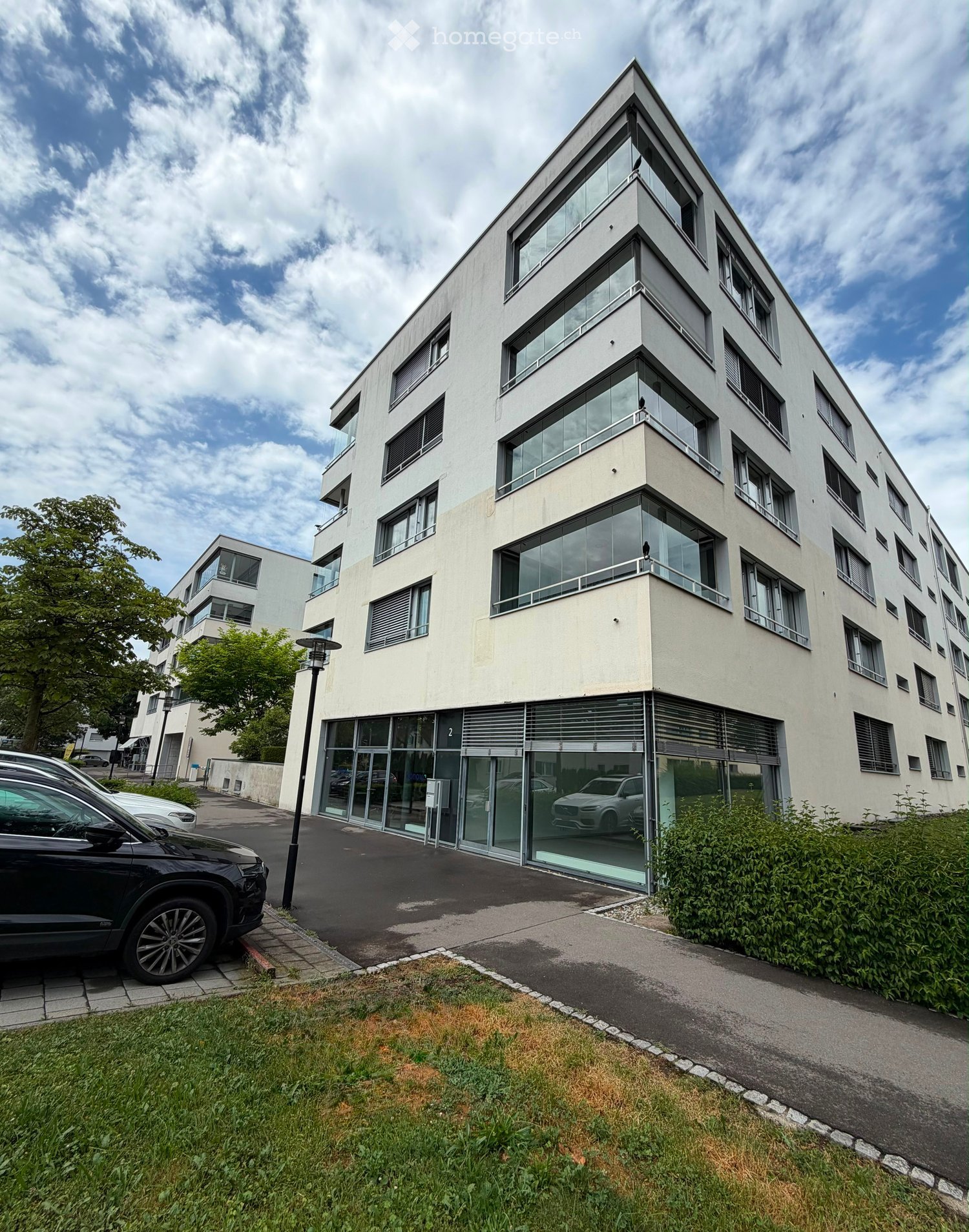 Modern multi-story building with large glass windows and balconies, surrounded by greenery and parked cars.