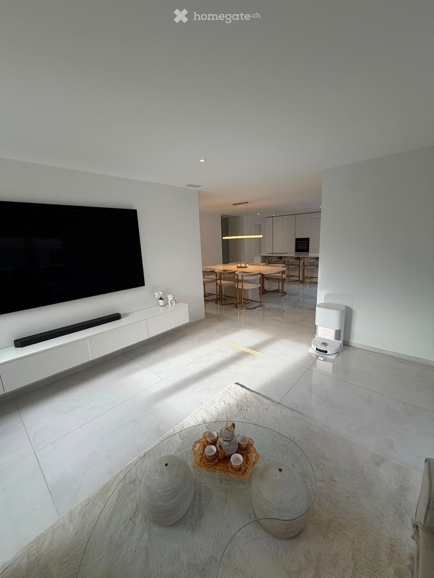 Modern living room with white walls, large TV, glass coffee table, decorative tea set, and a dining area with chairs and a table in the background.