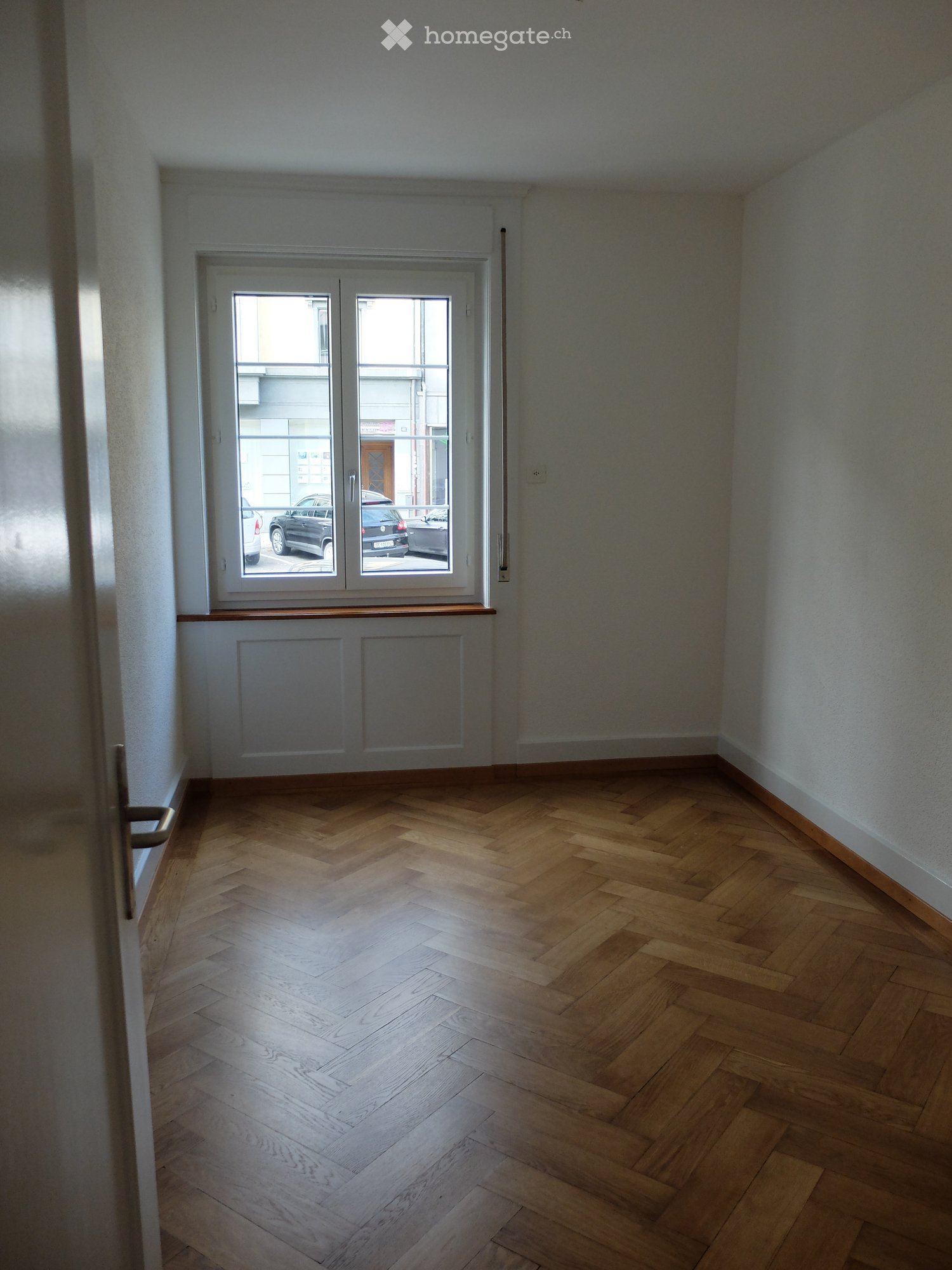 Empty room with wooden herringbone flooring, white walls, and a large window with double glass doors leading to a balcony.