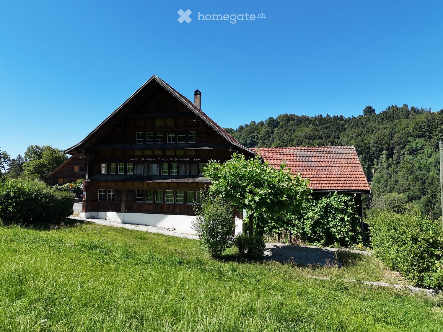 Traditional wooden house with a steeply pitched roof, surrounded by greenery and a small garden area.