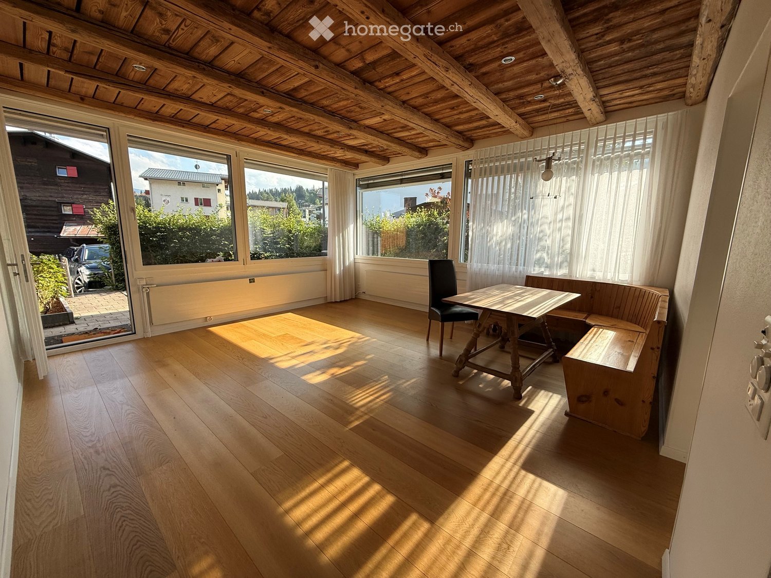 Bright room with wooden floor, large windows, a wooden table, a chair, and a bench. The room has a wooden ceiling with exposed beams and a view of the outdoors.