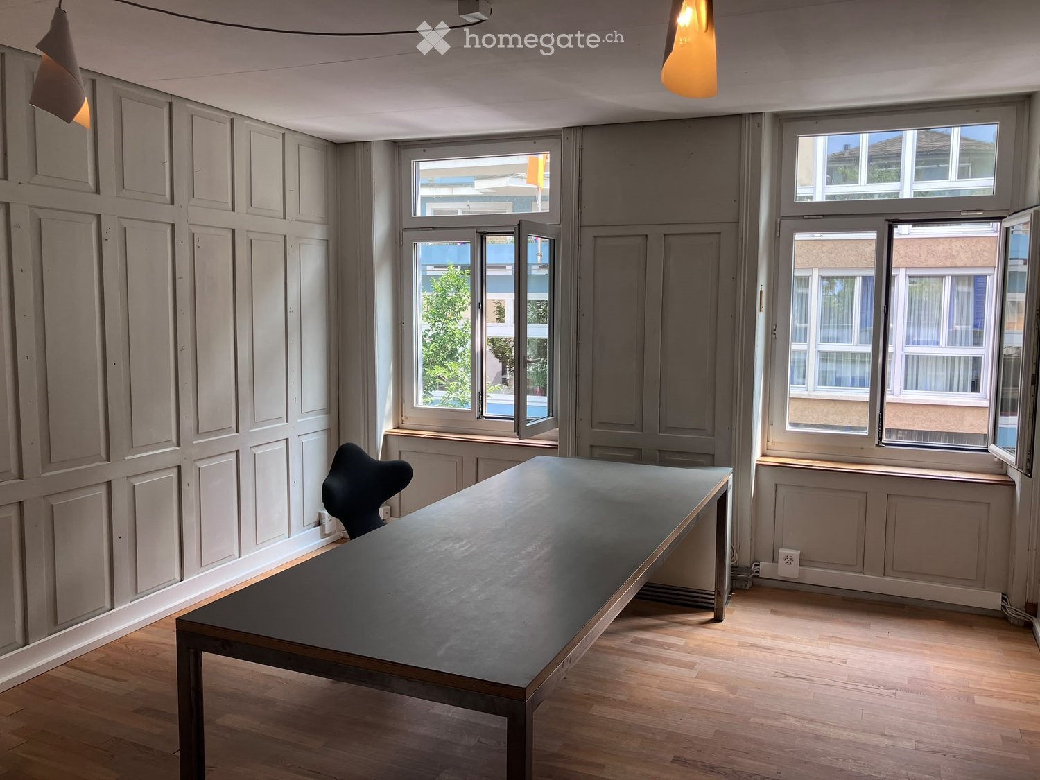 Minimalist room with wooden flooring, grey walls, large windows, a long dark table, and a single chair.