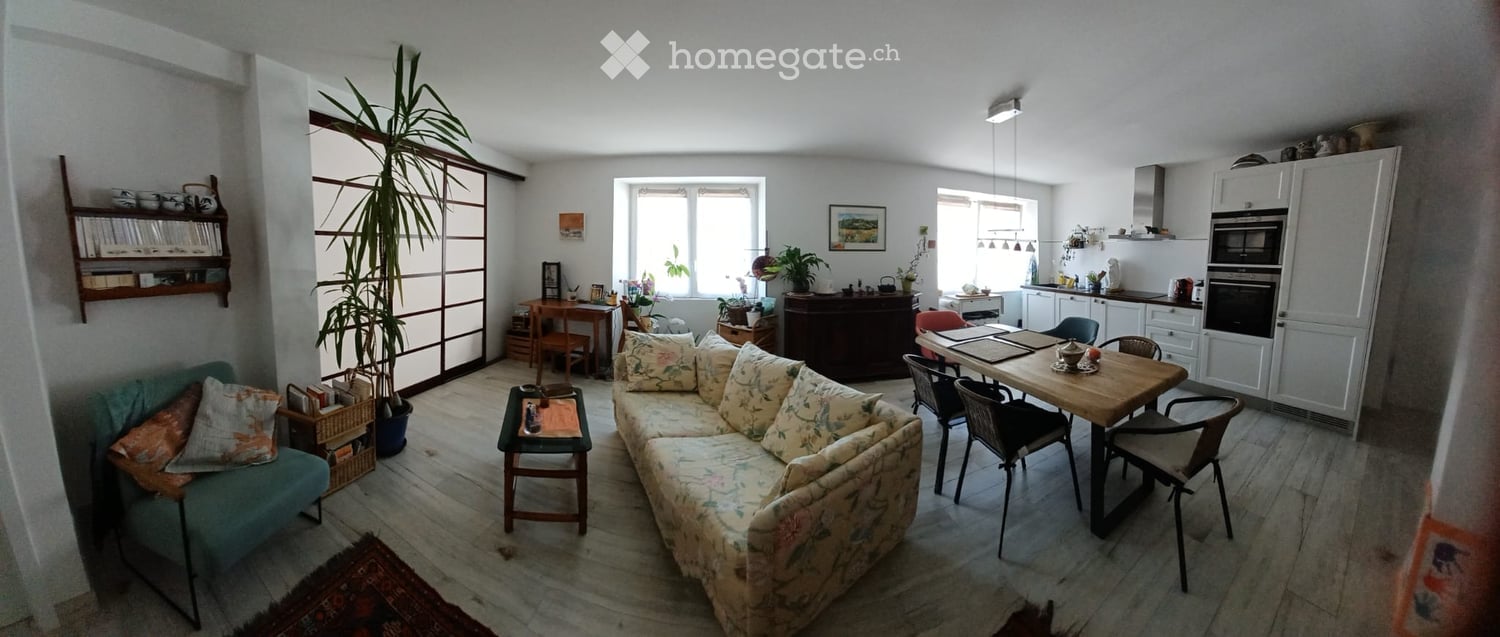 A well-lit living and dining area with a floral-patterned sofa, coffee table, dining table with chairs, and a kitchen area with white cabinets and appliances.