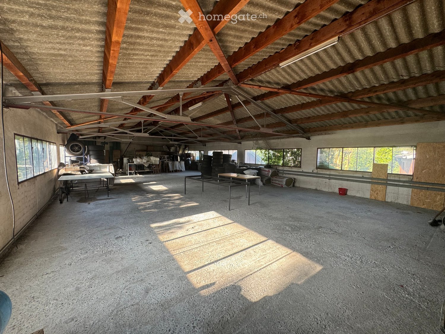 An empty, spacious room with a concrete floor and exposed wooden beams on the ceiling. The room has large windows allowing natural light to enter, and there are some storage items and equipment visible in the background.