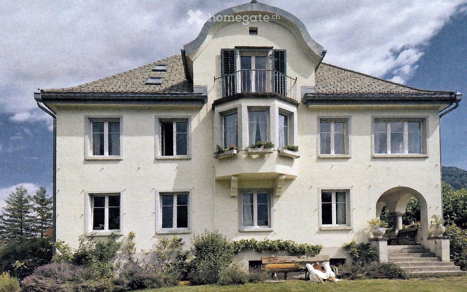 A large, elegant white house with multiple windows, balconies, and a curved roofline. The house features decorative window frames, potted plants on the balcony, and a well-maintained garden with a bench and steps leading to the entrance.