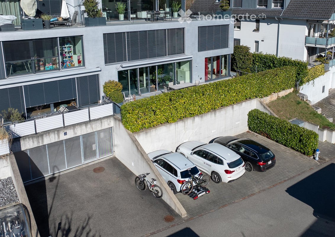 Modern multi-story residential building with large windows, balconies, and a parking area with several cars and bicycles.