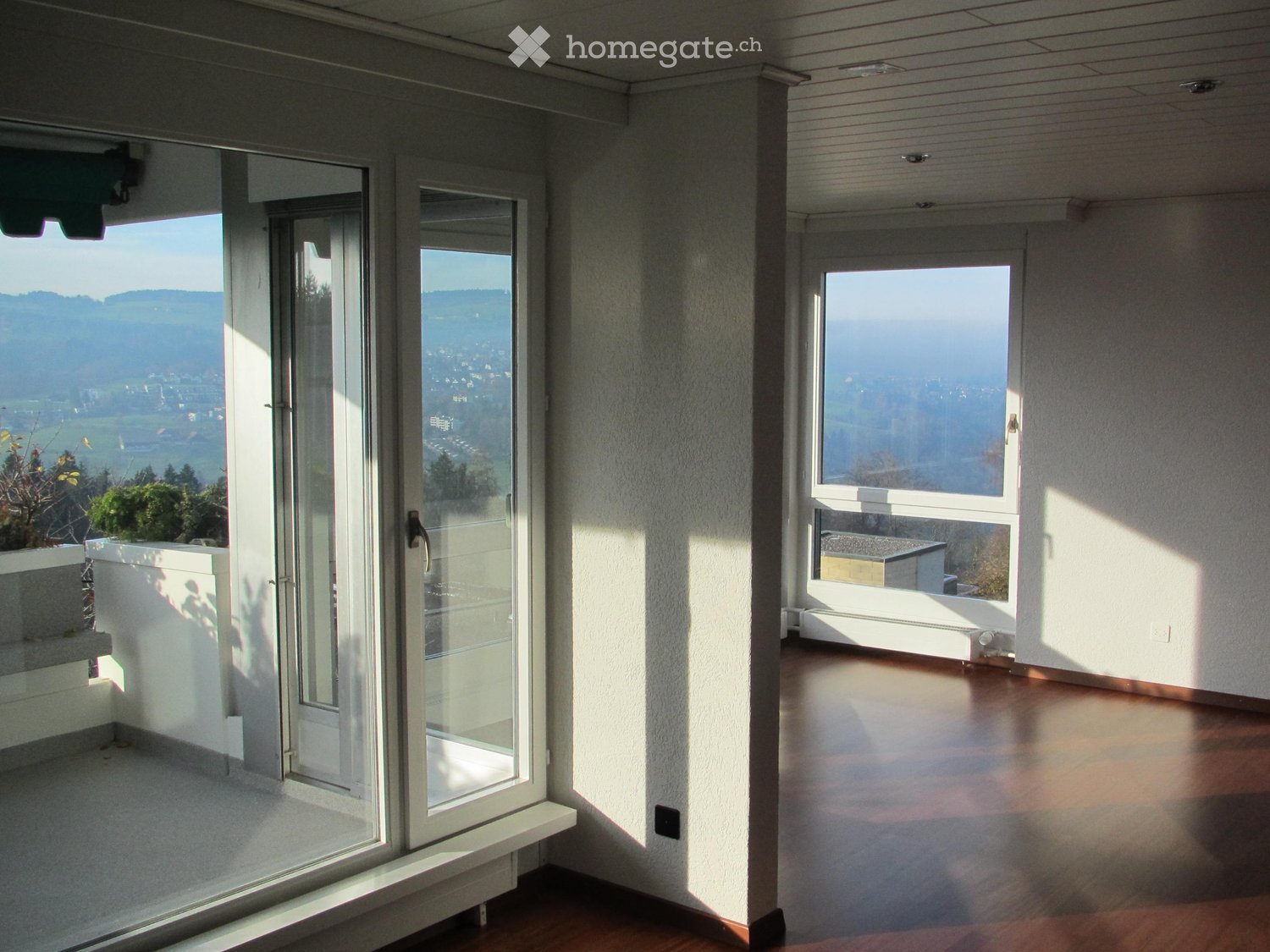 Bright, empty room with large windows and sliding glass doors leading to a balcony, offering scenic views of the landscape.