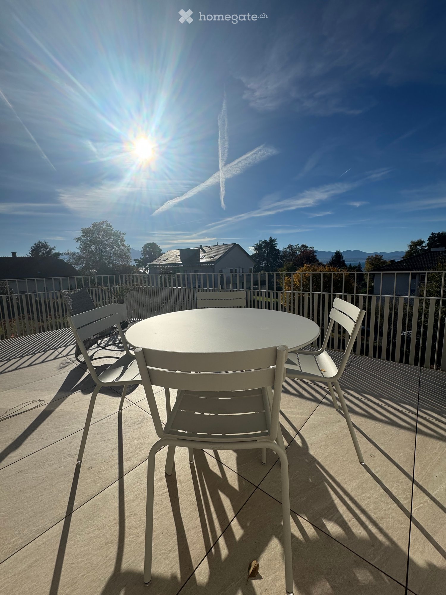 Balcony with a round white table and four white chairs, overlooking a residential area with trees and mountains in the background.