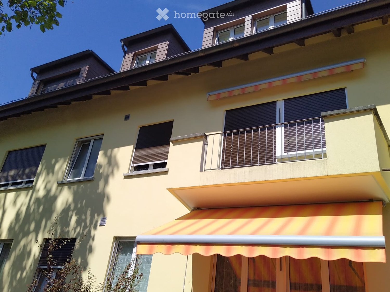 A multi-story residential building with yellow walls, multiple windows, balconies, and an awning over the entrance.