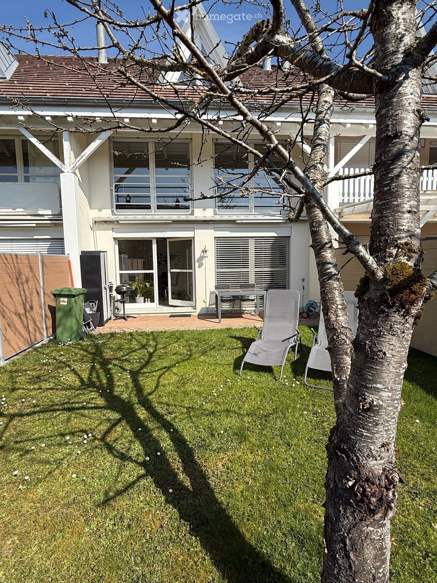 Well-maintained garden with green grass, a tree, and outdoor furniture including chairs and a table near the house entrance.