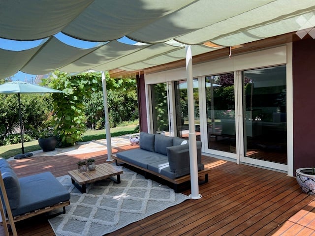 A modern outdoor terrace with wooden decking, grey sofa, coffee table, and a large umbrella providing shade. The space is connected to a house with large glass sliding doors.