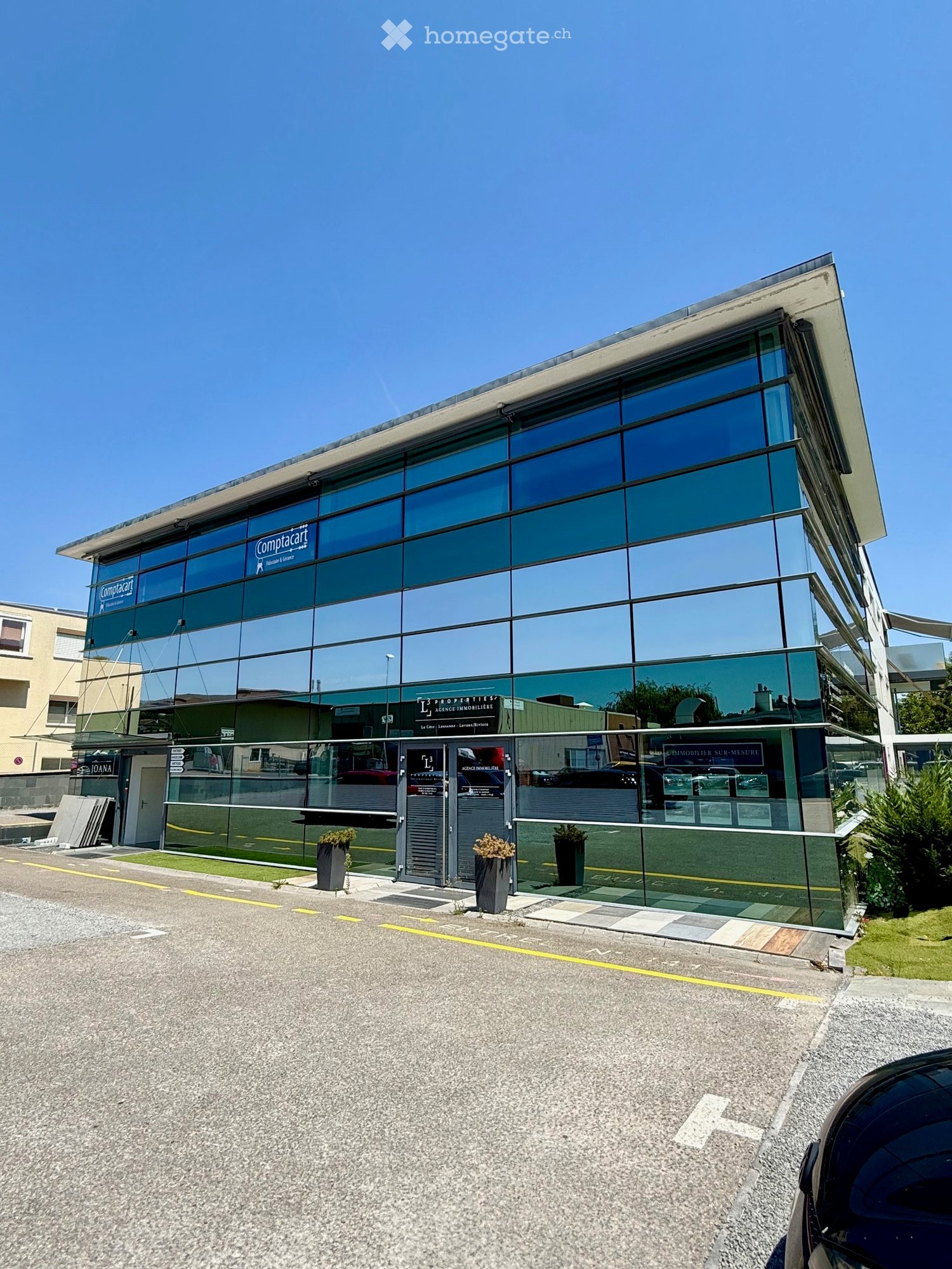 Modern commercial building with large glass windows and signage for 'Comptacart' and 'PROPERTIES' real estate agency. Features a clean, contemporary design with potted plants near the entrance.