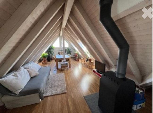 Cozy attic space with wooden beams, exposed wooden floor, grey sofa, coffee table, dining area with table and chairs, television, and a fireplace.