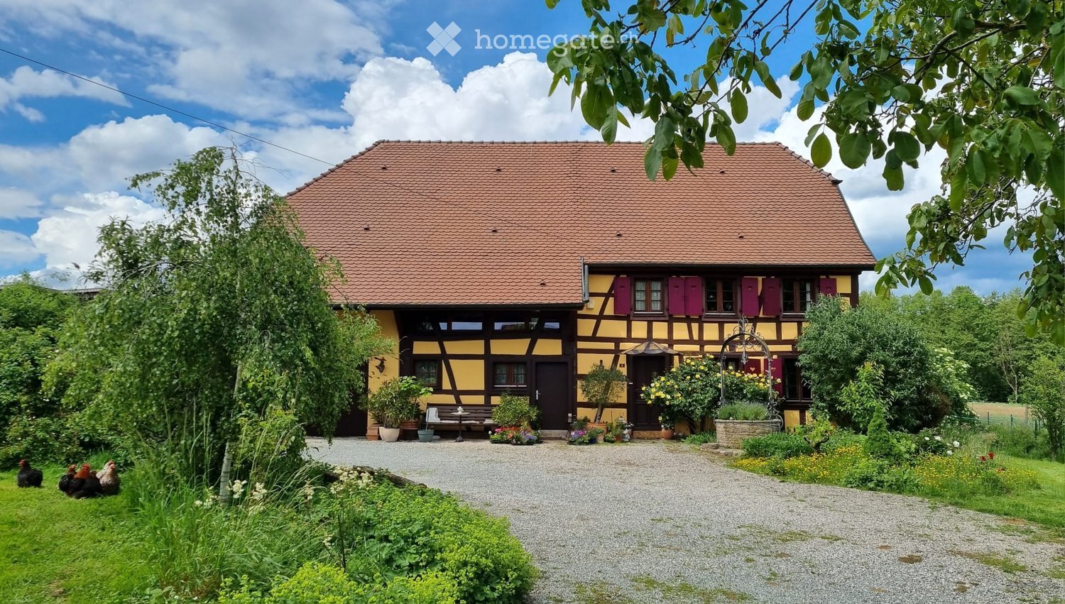 Traditional half-timbered house with yellow and brown exterior, red-tiled roof, surrounded by garden and gravel driveway.