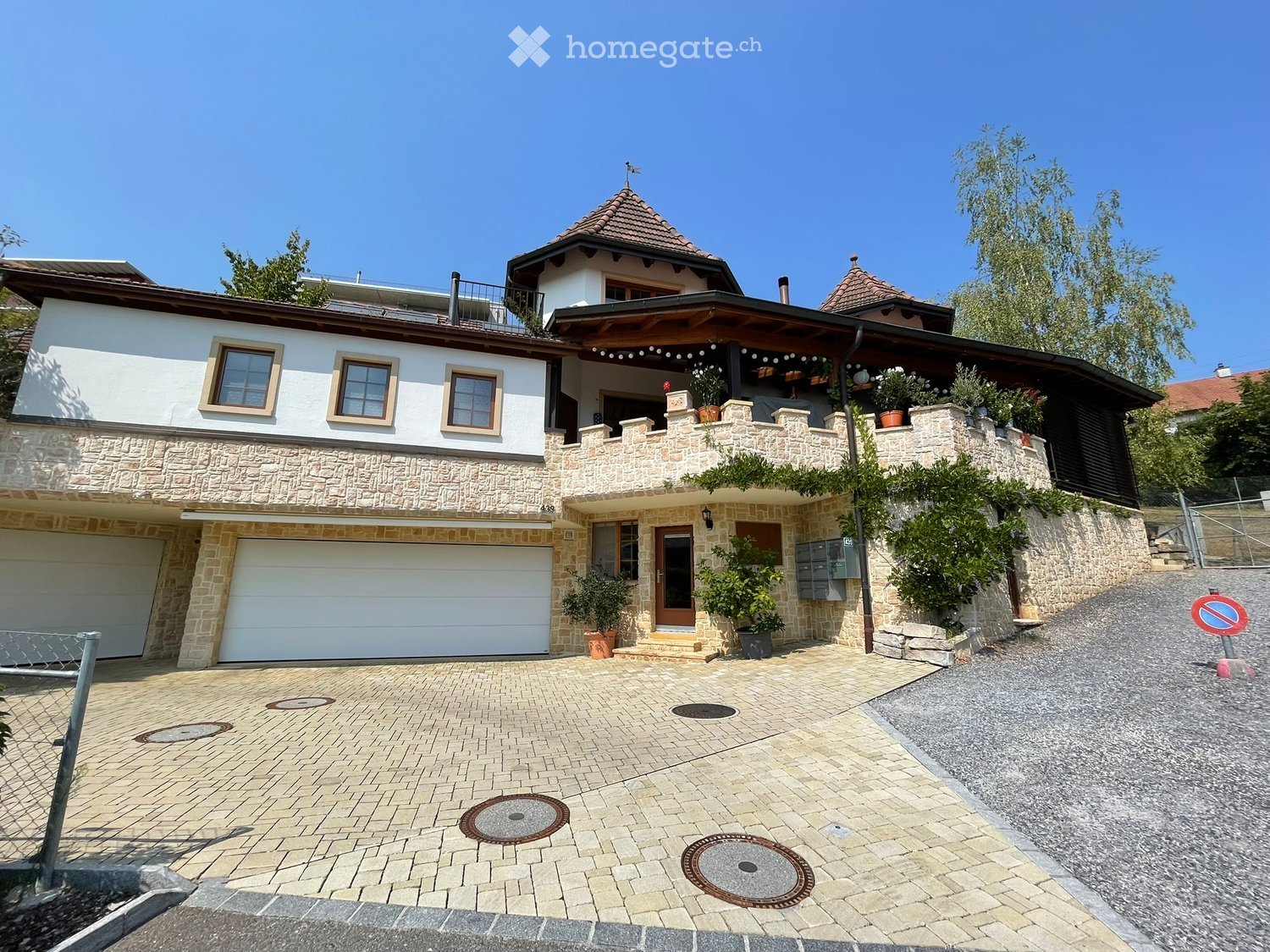Two-story house with a stone facade, garage, and balcony area with plants. The driveway is paved with light-colored bricks.