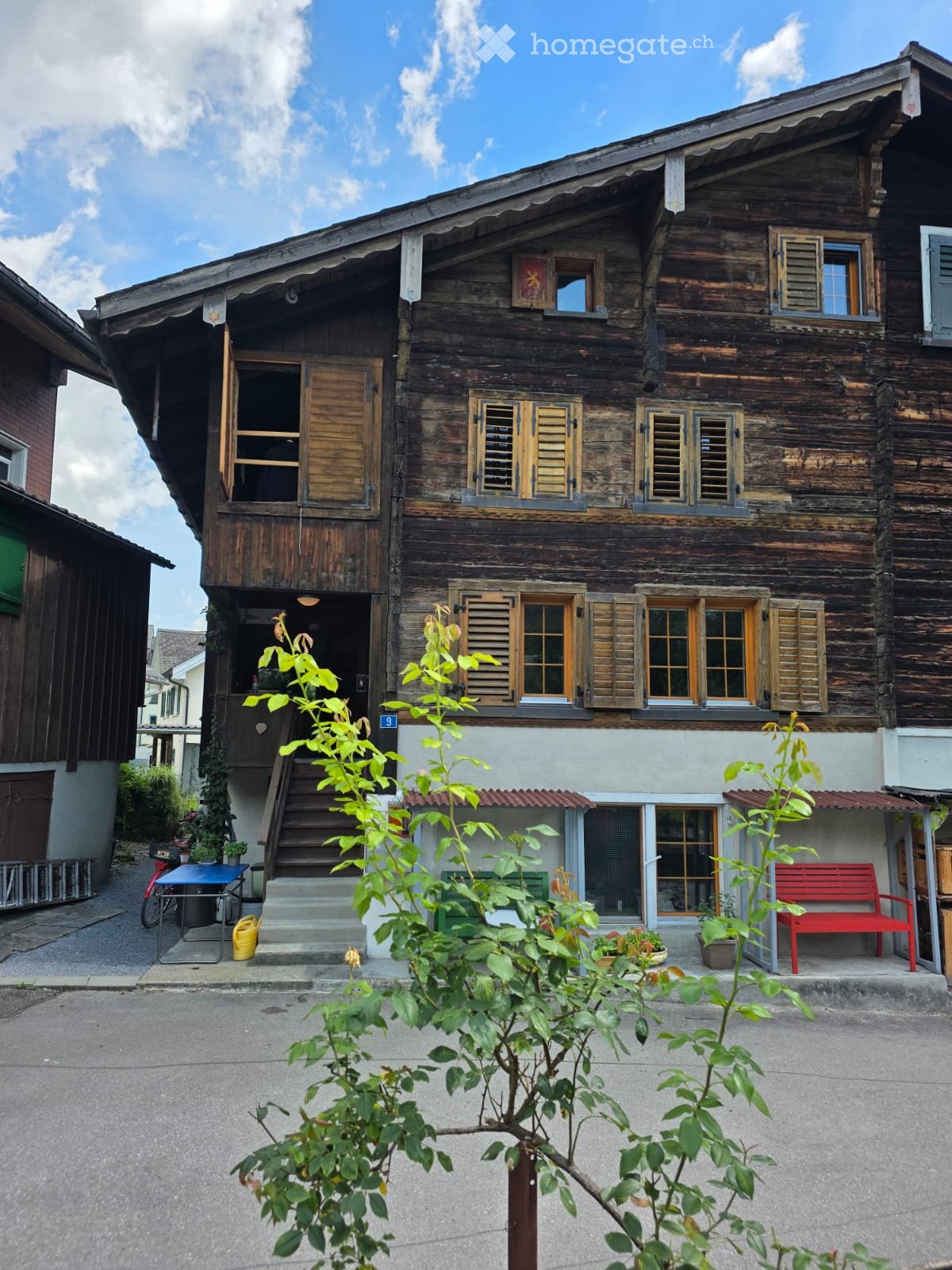 Traditional wooden house with multiple windows, shutters, and a small porch area with a red bench and plants.