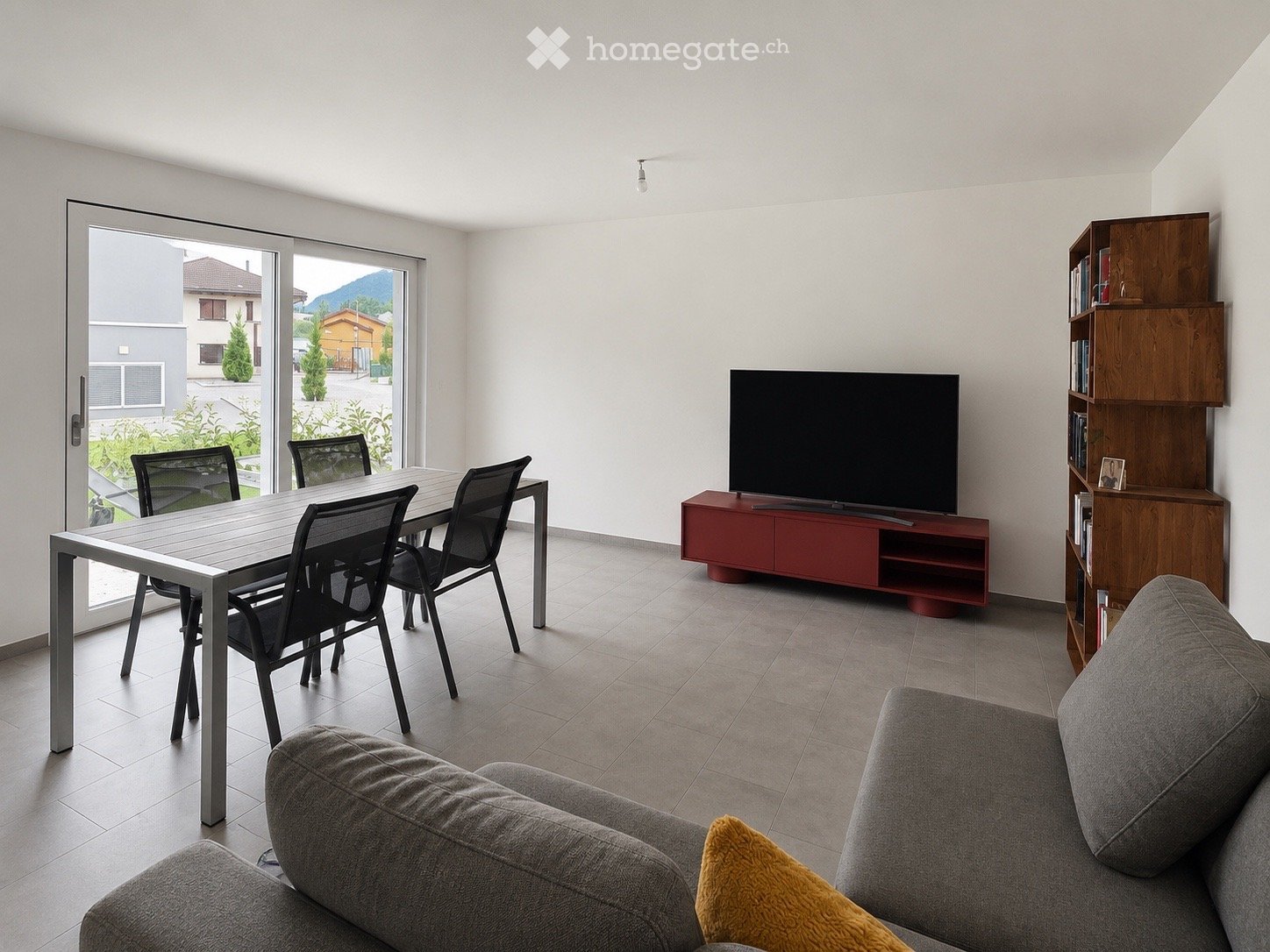 Modern living room with a grey sofa, a wooden shelf, a flat-screen TV on a red stand, a dining table with four chairs, and a large window with a view of the outdoor area.