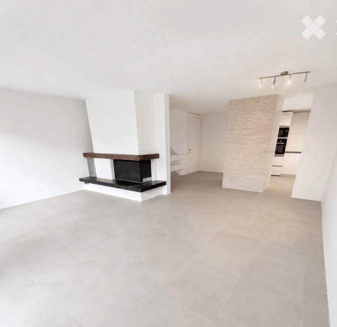 Open-plan living space with a fireplace, white walls, grey tiled floor, and recessed lighting. The room is unfurnished, showing only the fireplace and a doorway leading to another area.