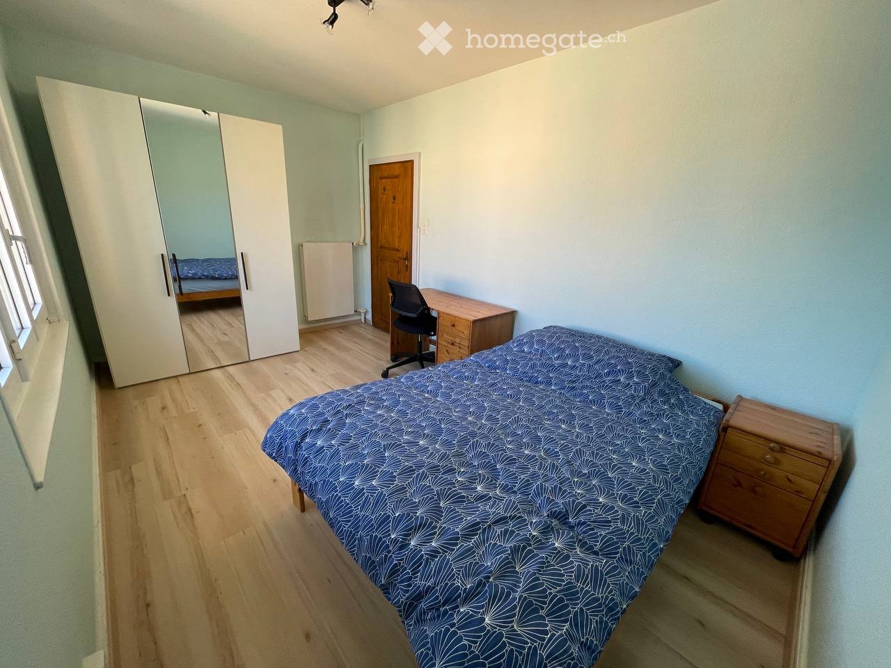 Cozy bedroom with light blue walls, wooden flooring, a single bed with blue patterned bedding, a wooden nightstand, a large mirrored wardrobe, a desk with a chair, and a radiator.