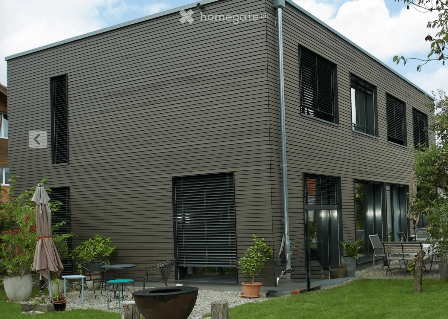 Modern house exterior with grey wooden siding, large windows, and a well-maintained garden area featuring potted plants, outdoor furniture, and a fire pit.