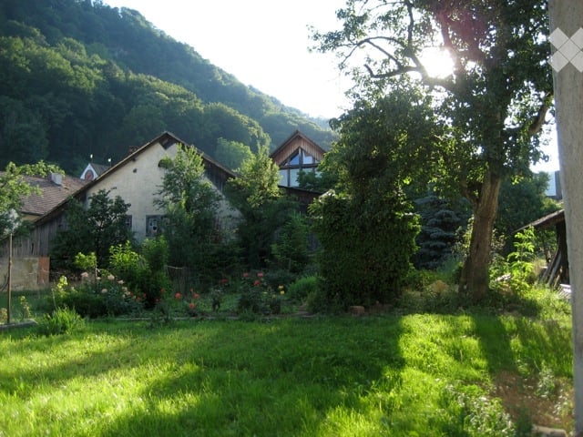 A peaceful garden area with a well-maintained lawn, surrounded by trees and shrubs. A small house with a stone facade and wooden roof is visible in the background, nestled among greenery and hills.