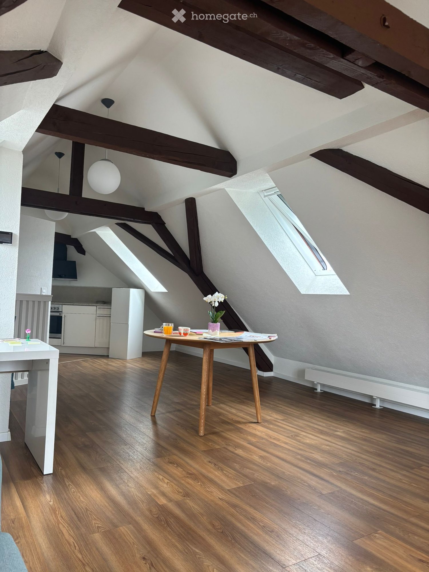 Bright attic room with wooden beams, wooden floor, skylight windows, small round table with chairs, and a kitchen area with white cabinets and appliances.