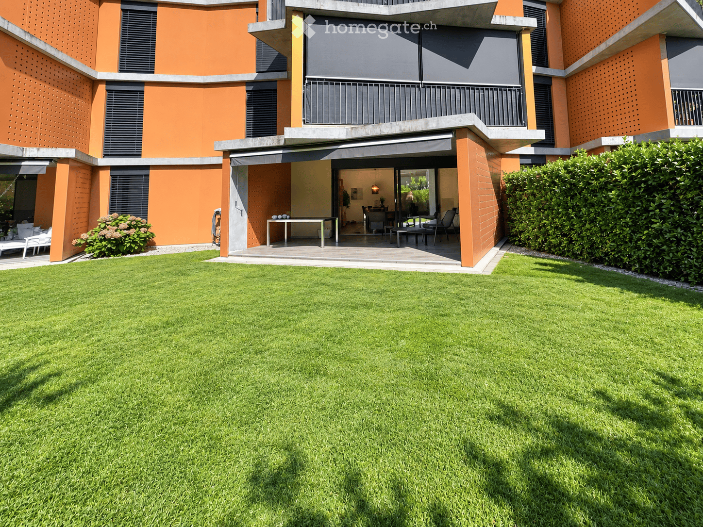 Modern house with orange and grey facade, large lawn, patio with outdoor furniture, and covered entrance area.