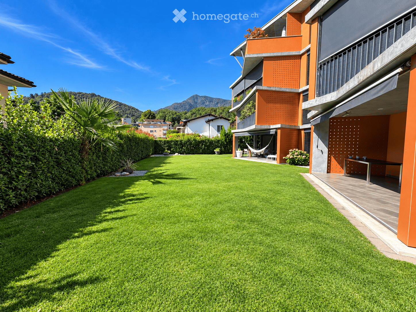 Well-maintained lawn with trimmed hedges, a modern building with orange accents, and a hammock on the patio.
