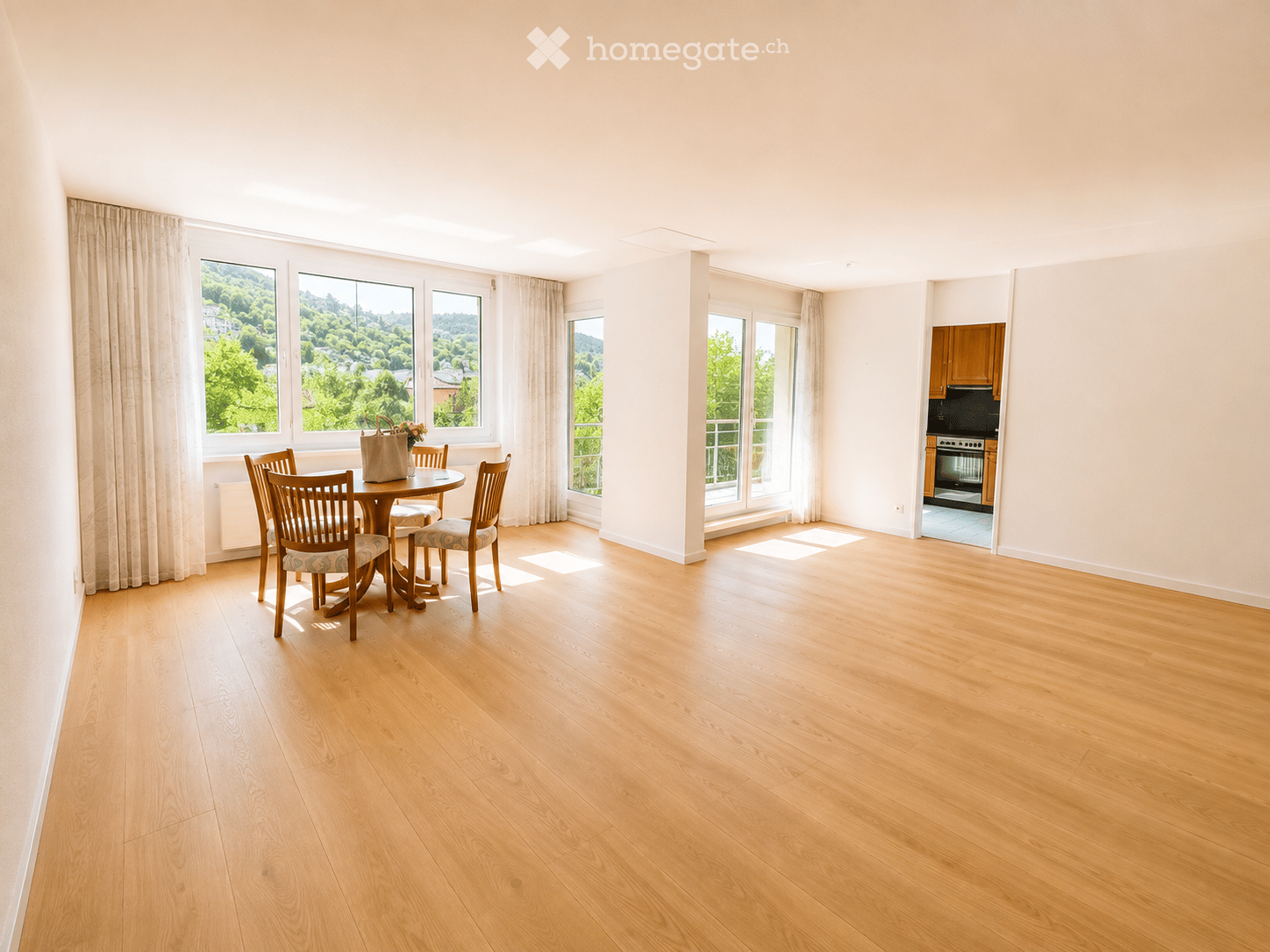 Bright, spacious room with wooden flooring, large windows with curtains, a dining table with chairs, and a handbag with flowers on it. A kitchen area is visible in the background.