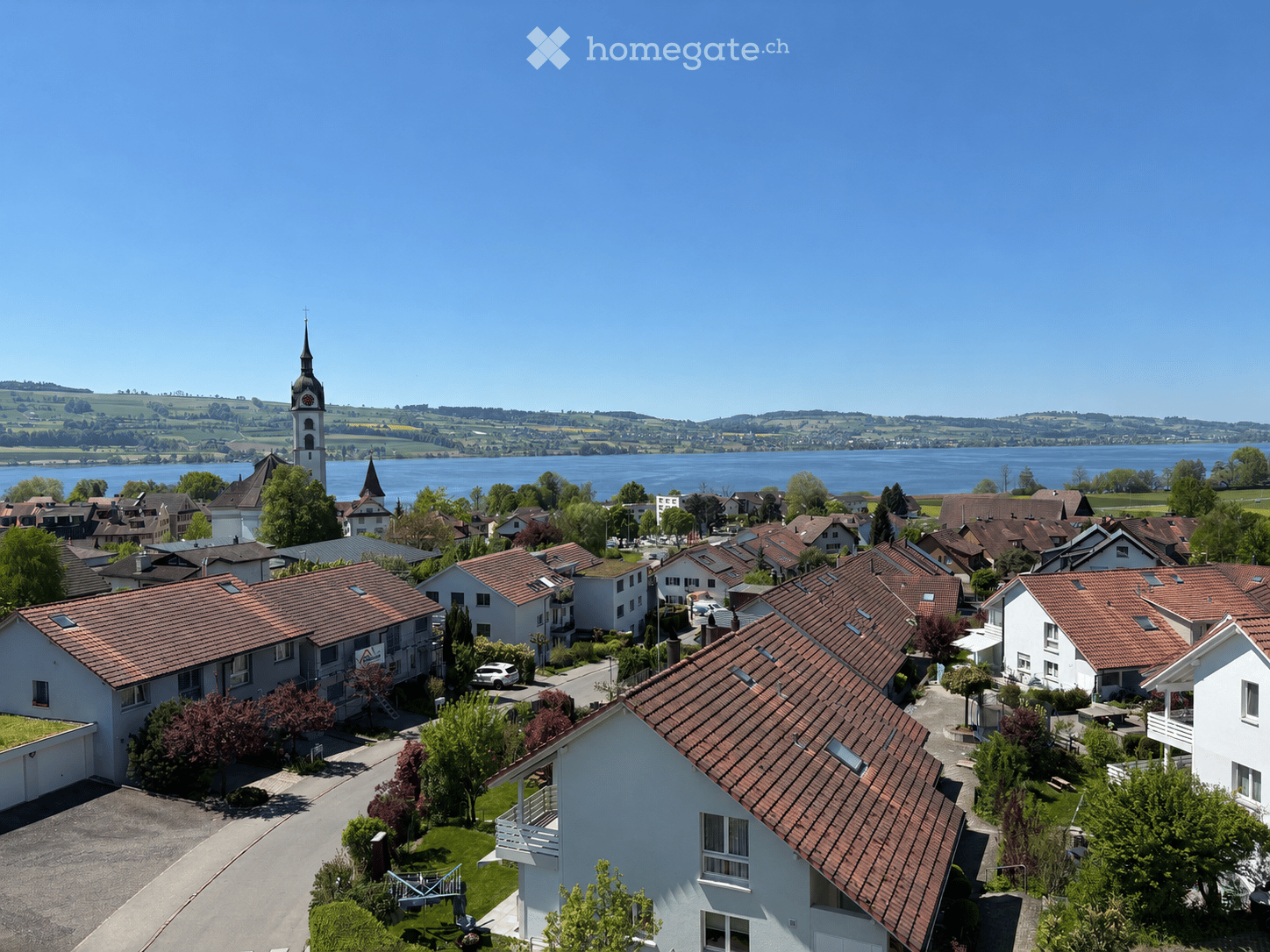 Aerial view of a residential area with multiple houses, red-tiled roofs, and a church tower overlooking a lake in the background.