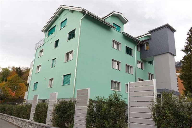 A modern residential building with three floors and several windows. The walls are painted green, and the roof has a triangular shape. The exterior is adorned with multiple white railings on the balcony, and there are bushes and trees on the sides. The bu