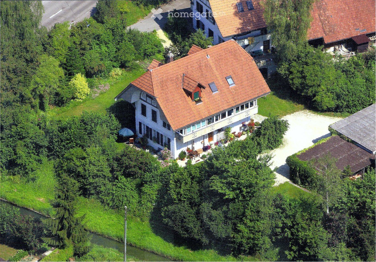 Aerial view of a house with a red roof, surrounded by trees and plants, with a driveway leading to it.