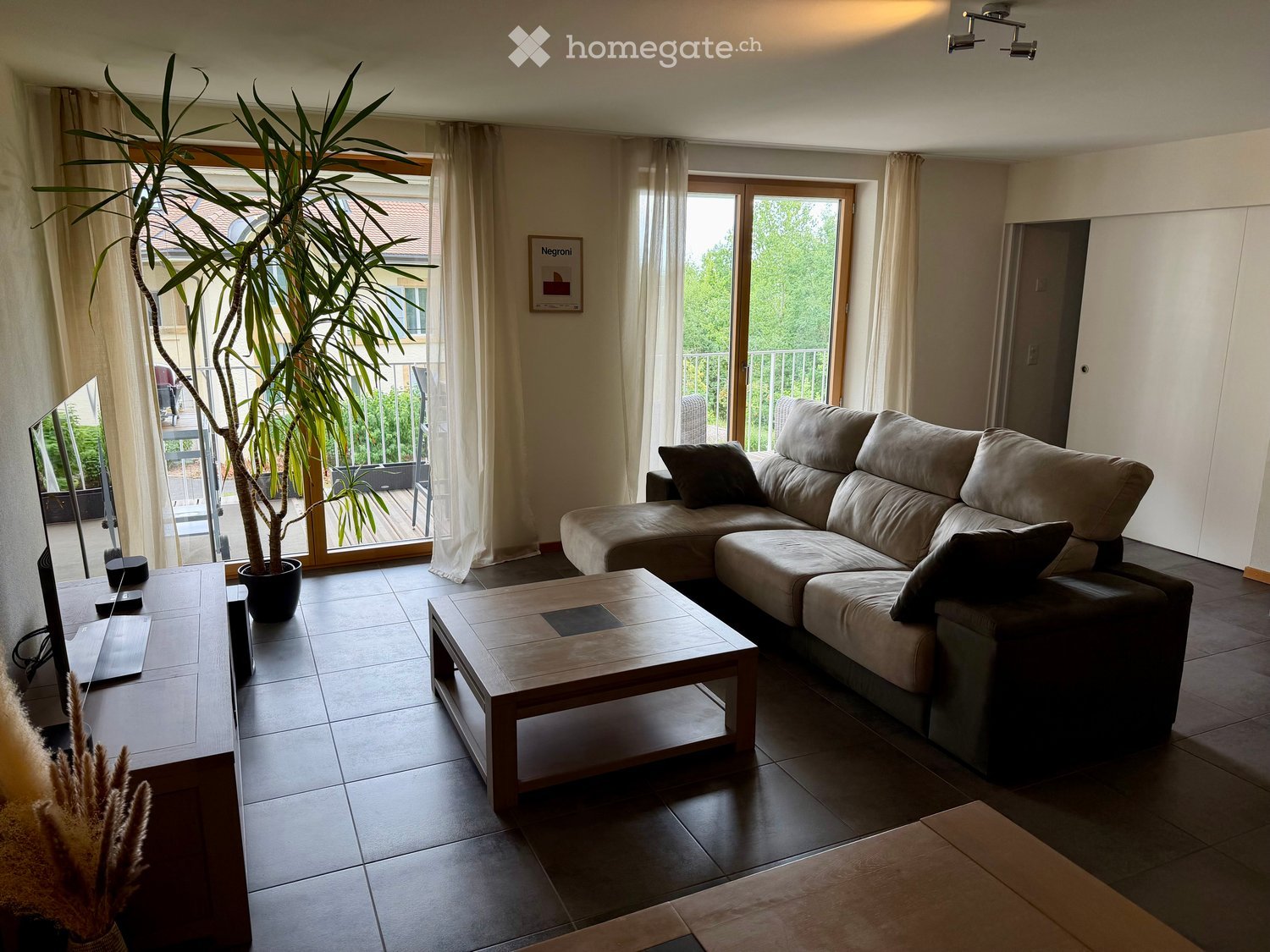 Modern living room with a large sectional sofa, wooden coffee table, TV stand, laptop, and a tall potted plant near the sliding glass doors leading to a balcony.