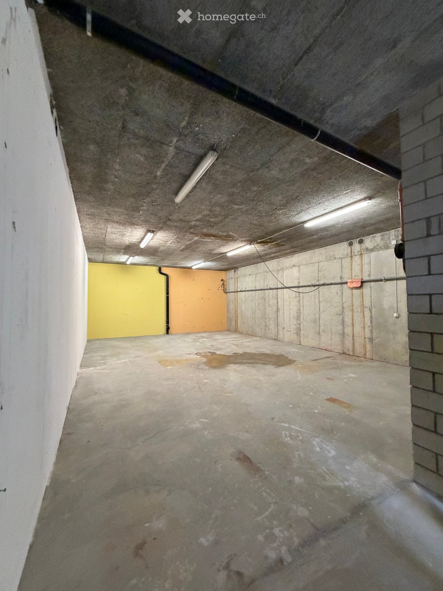 Empty underground parking space with concrete walls, ceiling, and floor. Features fluorescent lighting and a small yellow door at the far end.
