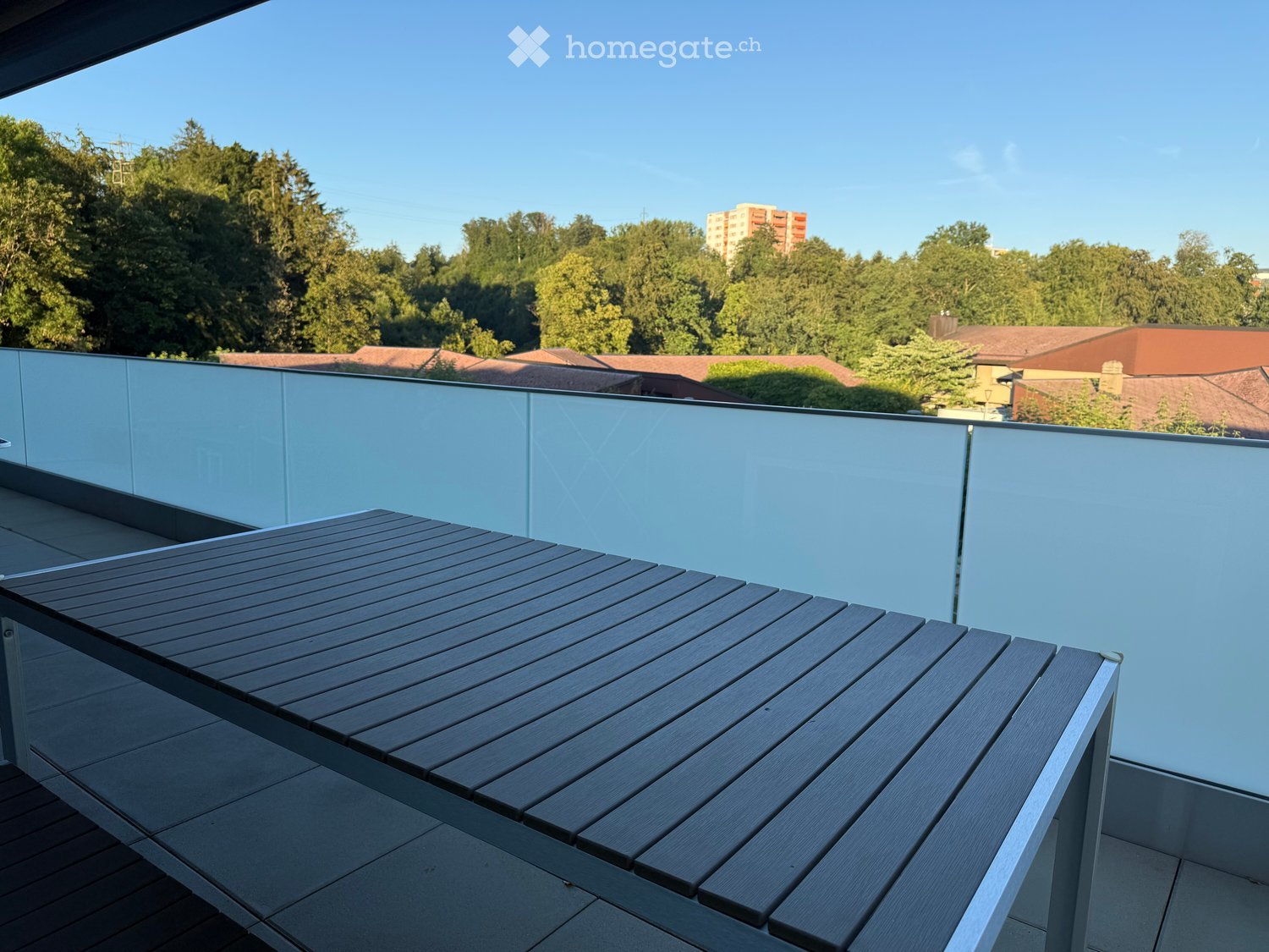 Modern balcony with a wooden table, glass railing, and a view of trees and buildings.