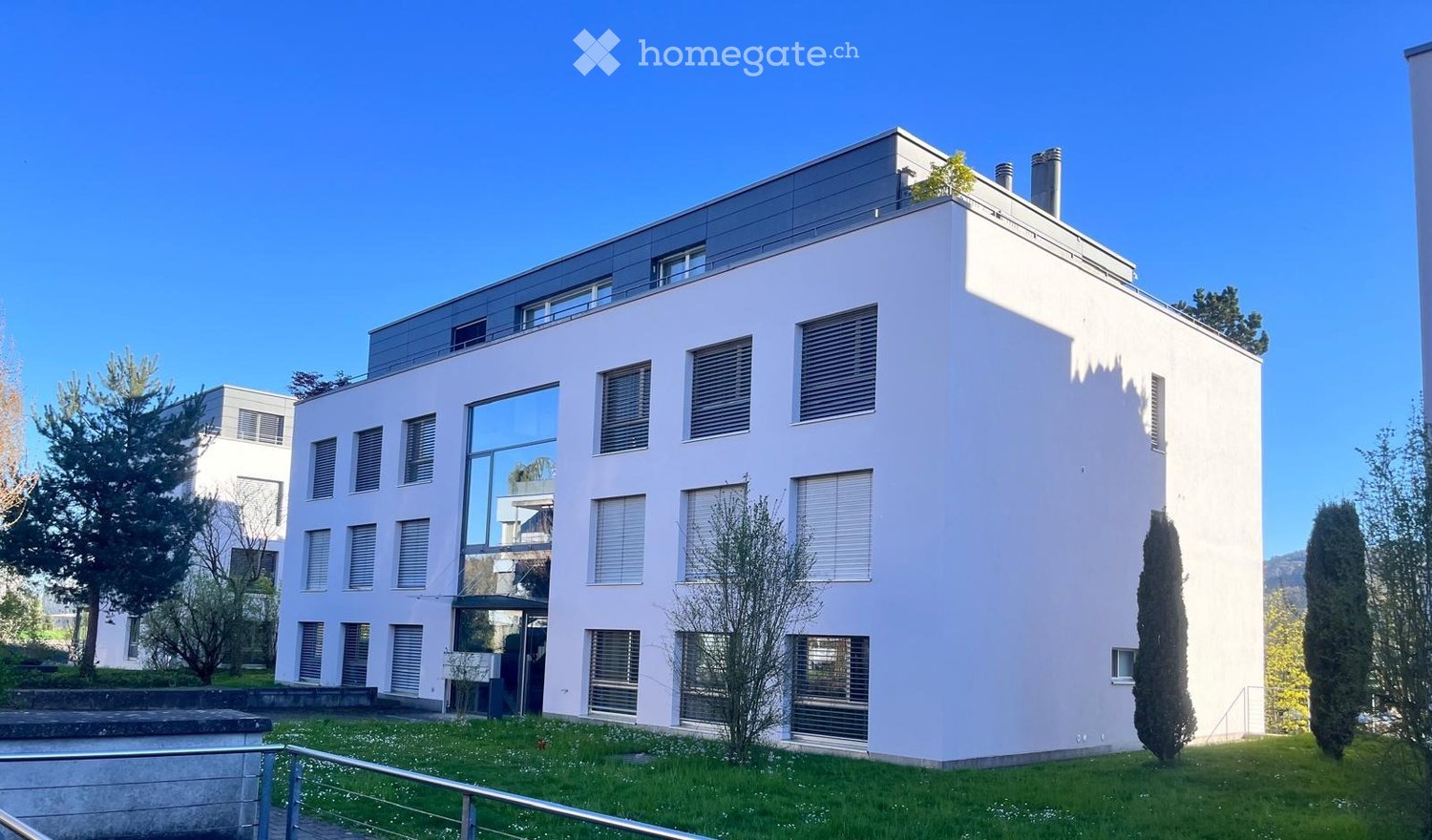 Modern white building with glass windows, multiple floors, surrounded by greenery
