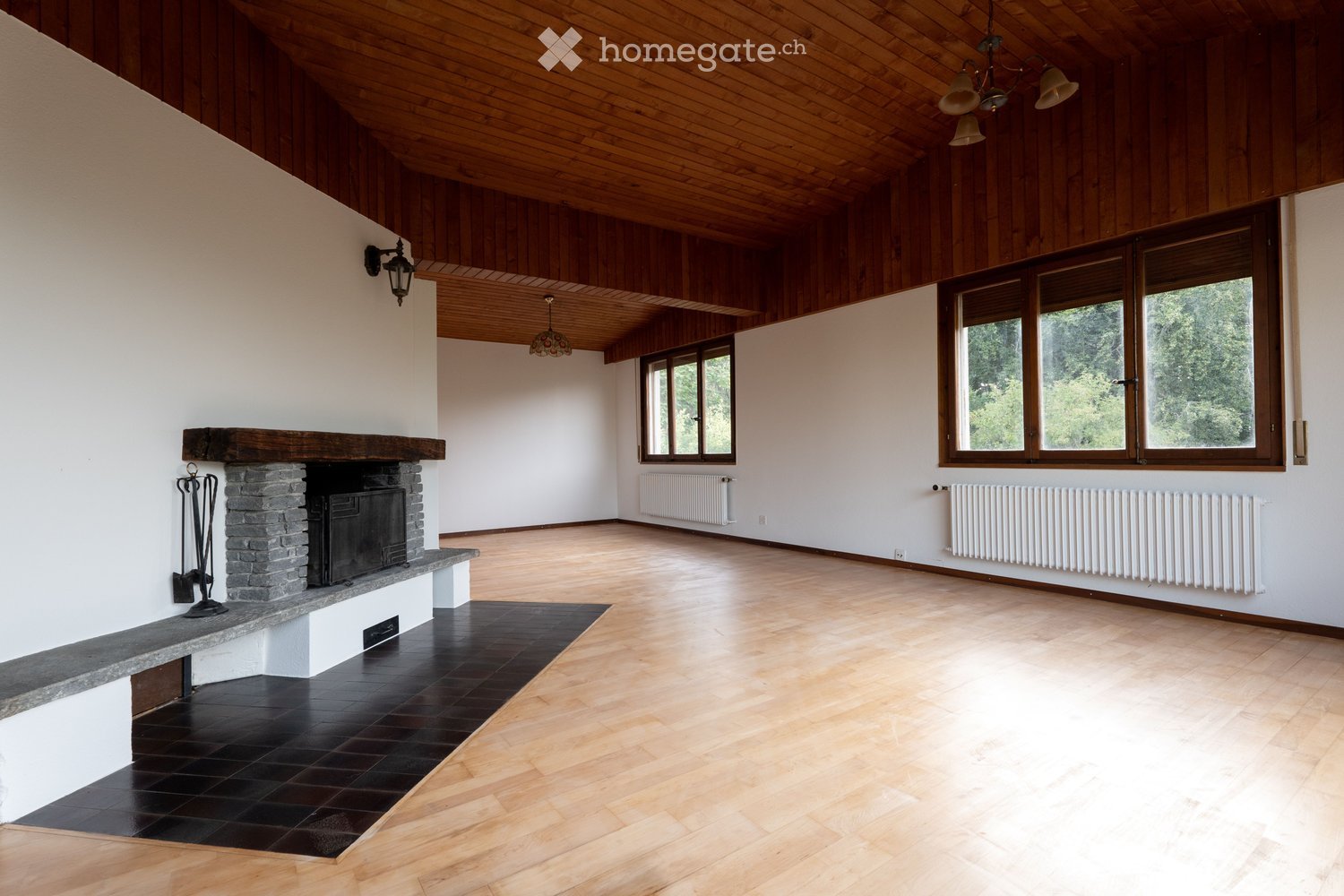 Spacious, unfurnished room with wooden floors, white walls, large windows, and a stone fireplace. The room features exposed wooden beams on the ceiling and radiators along the walls.