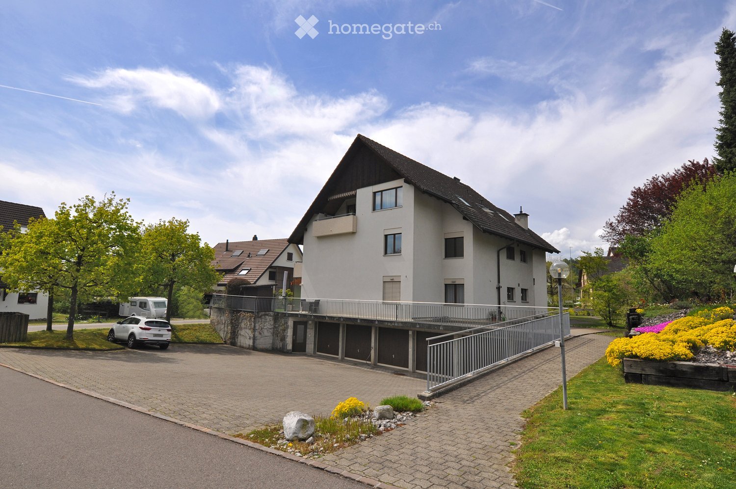 Two-story house with white exterior, dark roof, and balcony on the upper level. The house is surrounded by a well-maintained garden with colorful flowers and trees. There is a paved driveway leading to a garage area with multiple parking spaces.