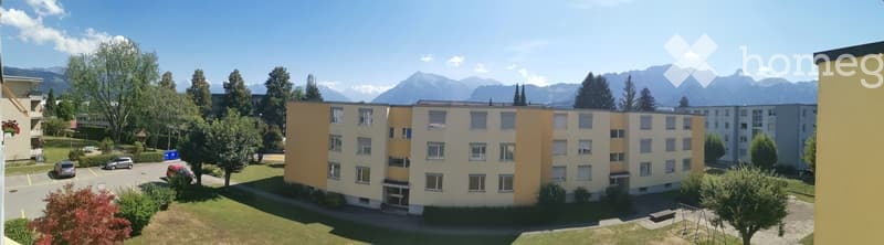 A multi-story residential building with light yellow and cream-colored facades, surrounded by trees and parked cars, with a scenic mountain backdrop.