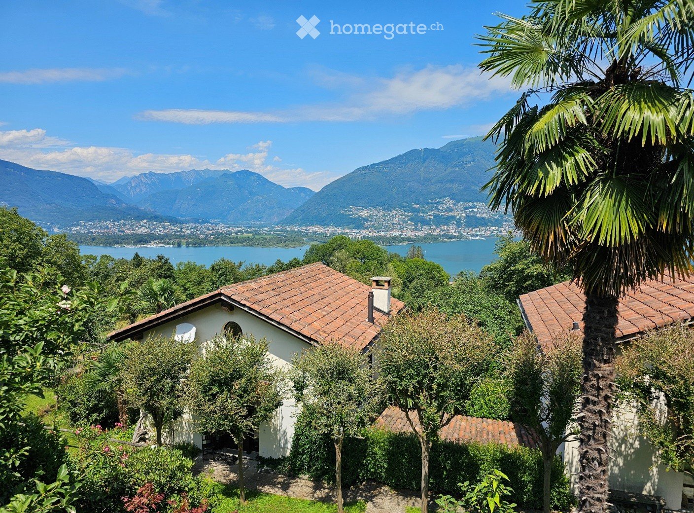 A serene garden view with a small white house, surrounded by lush greenery and palm trees, overlooking a lake and distant mountains.